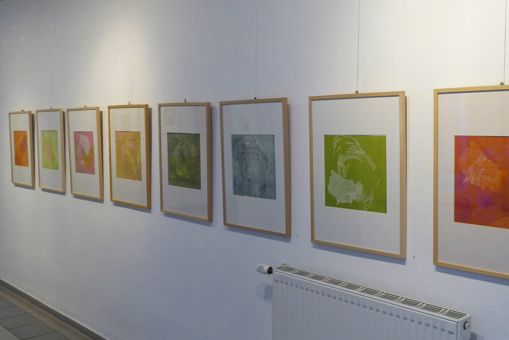 Ausstellungsansicht mit acht sehr unterschiedlichen Varianten aus der Serie von farbigen Holzschnitten und Monotypien zum Thema "Spuren Weitergesponnen."
Die Holzschnitte sind teils stark grafisch in kräftig bunten Farben Pink mit Orange mit weiß, teils grün-weiß und teils sehr zahrt malerisch zum Beispiel in hellgrün, rosa und gelb oder nur in hellem blau-grau